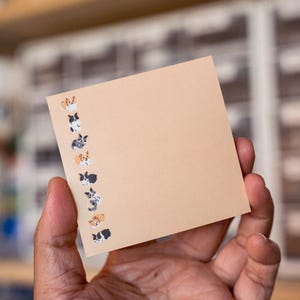 May include: A square, light tan sticky note featuring a vertical column of small, colourful illustrations of various dogs along the left edge. The dogs are depicted in different poses and colour patterns, including black, white, and brown markings.