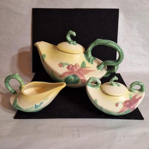 May include: A three-piece ceramic tea set. The set includes a teapot, a creamer, and a sugar bowl. The pieces are cream-colored with yellow tops, green handles, and pink and green floral designs. The teapot measures approximately 8 inches in length.