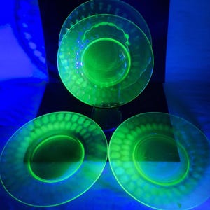 May include: Three circular glass plates glowing with a bright neon green hue under ultraviolet light. Each plate features a smooth center surrounded by a textured rim with a repeating pattern of small, rounded indentations along the outer edge.