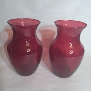 May include: Two matching ruby-colored glass vases. Each vase has a rounded body and a flared rim. The glass has a glossy finish, reflecting light. The vases are suitable for displaying flowers or as decorative pieces.