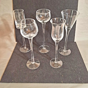 May include: Five clear glass stemware glasses of varying shapes and sizes are arranged on a dark surface. The glasses include a shot glass, a wine glass, a cordial glass, a tulip-shaped glass, and a martini glass.