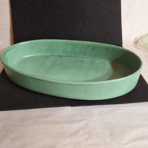 May include: An oval-shaped ceramic baking dish with a matte, light green finish. The interior surface features a subtle, mottled texture with darker green speckling throughout, while the exterior walls are smooth and consistent in color.