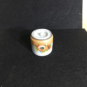 May include: A small, white ceramic jar with a lid. The jar has a brown band around the middle and is decorated with orange flowers.