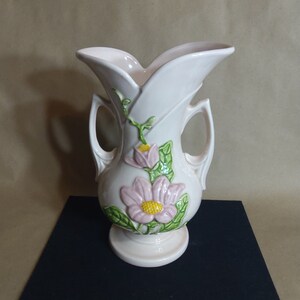May include: A pink ceramic vase with two handles and a floral design. The vase features a pink flower with green leaves and a yellow center.
