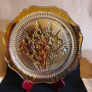 May include: An amber-colored glass plate with a scalloped edge. The plate features a raised floral design in the center. The plate is supported by a red wooden stand. The plate has a textured surface.