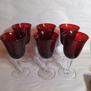 May include: Six wine glasses with a clear stem and base, featuring a deep red bowl. The glasses have a flared rim and a dark burgundy exterior. The glasses are arranged on a white surface.