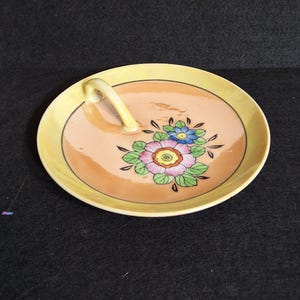 May include: A round ceramic dish with a yellow rim and a peach-colored center, featuring a single arched handle. The surface is decorated with a painted floral motif in shades of pink, blue, green, and yellow, with black outlines and details.