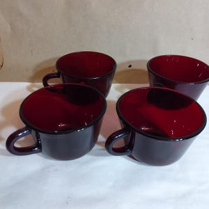May include: Four burgundy-colored coffee cups with black rims and handles. The cups have a glossy finish and a deep, rich color. The cups are arranged in a group, suggesting a set for serving beverages.