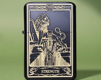 May include: A matte black metal lighter featuring a gold-toned engraved design of a tarot card. The graphic depicts a lion and a skeleton figure with the Roman numeral VIII at the top and the word STRENGTH printed at the bottom of the frame.