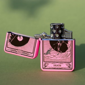 May include: Pink lighter with a silver top and a black and white image of the Death tarot card. The card features a skeleton holding a scythe and the word "DEATH" is printed below the image.