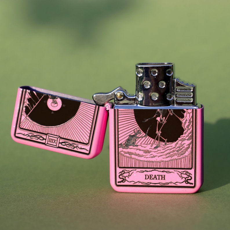 May include: Pink lighter with a silver top and a black and white image of the Death tarot card. The card features a skeleton holding a scythe and the word "DEATH" is printed below the image.