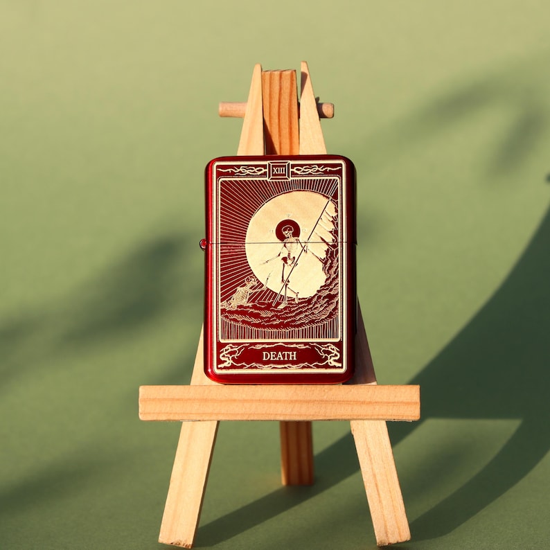 May include: A red lighter with a gold design of the Death tarot card. The card features a skeletal figure holding a scythe and a sun behind it. The lighter is displayed on a wooden easel.