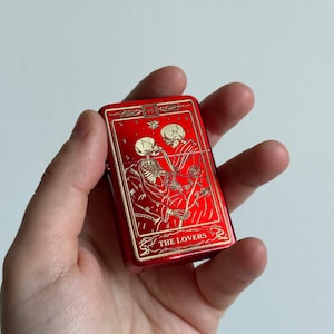 May include: Red metal lighter with a gold design of two skeletons embracing. The design includes the text "THE LOVERS" and the Roman numeral "VI".