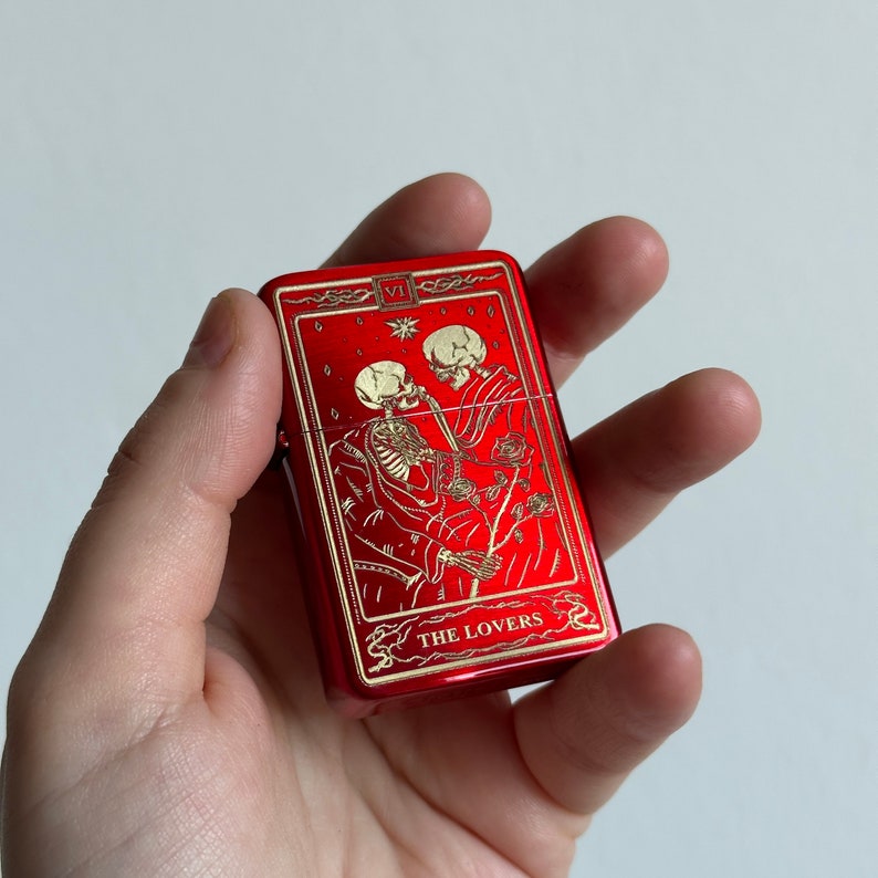 May include: Red metal lighter with a gold design of two skeletons embracing. The design includes the text "THE LOVERS" and the Roman numeral "VI".