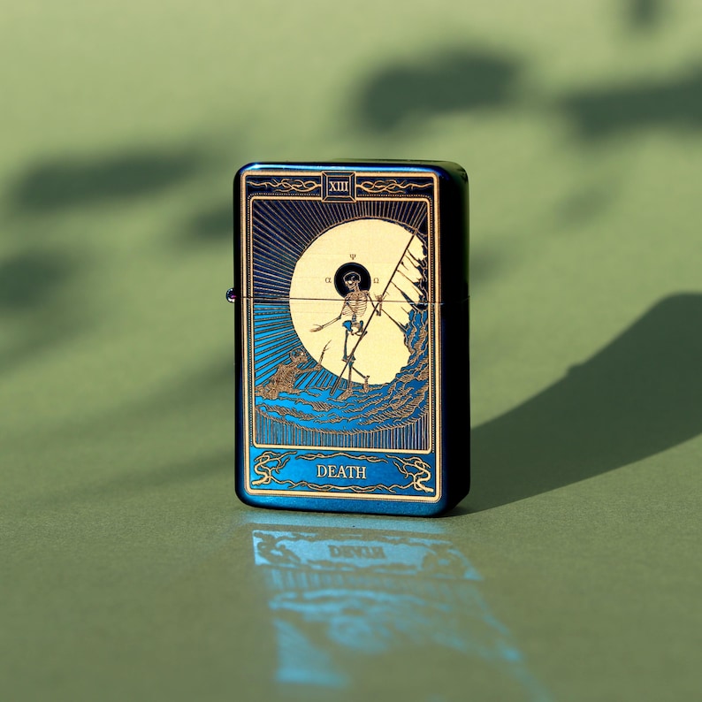 May include: A blue and gold lighter with a design of the Death tarot card. The card features a skeleton holding a scythe and a crescent moon.