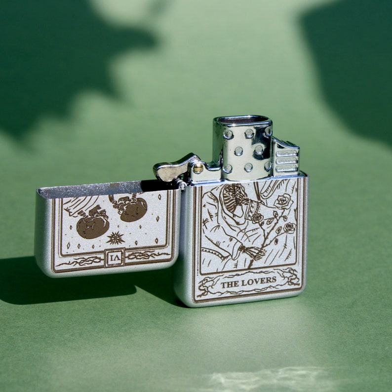 May include: A silver lighter with a design of the Lovers tarot card. The lighter is open and the design is visible on both the front and back of the lighter.