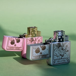 May include: Two silver and pink lighters with a design of playing cards. The lighters are open and show the card designs. The silver lighter has the card "The Lovers" on it. The pink lighter has a card with two hearts on it.