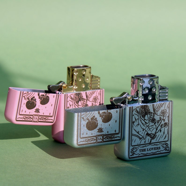 May include: Two silver and pink lighters with a design of playing cards. The lighters are open and show the card designs. The silver lighter has the card "The Lovers" on it. The pink lighter has a card with two hearts on it.