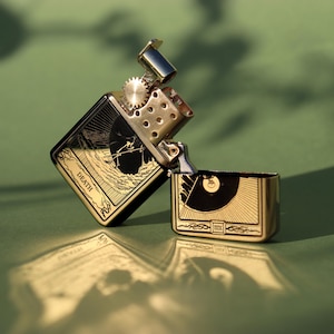 May include: A gold and black lighter with an engraved design of a skeletal figure holding a scythe. The lighter is open and the inscription "DEATH" is visible on the side.