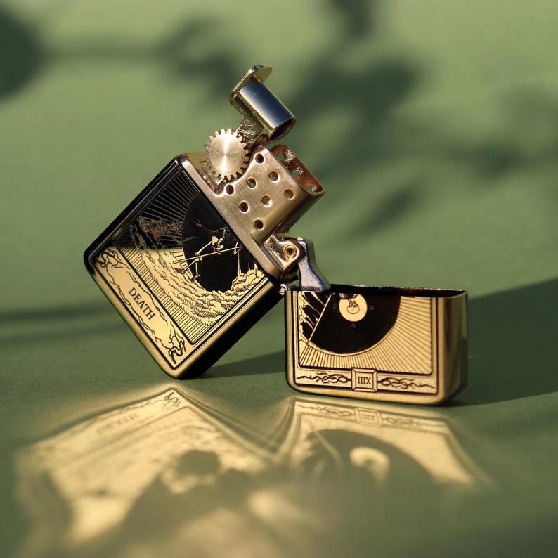 May include: A gold and black lighter with an engraved design of a skeletal figure holding a scythe. The lighter is open and the inscription "DEATH" is visible on the side.