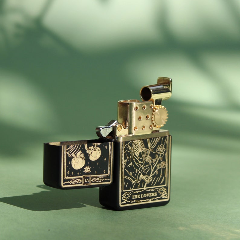 May include: A black lighter with gold accents and a design of the Lovers tarot card. The lighter is open and the flint wheel is visible.