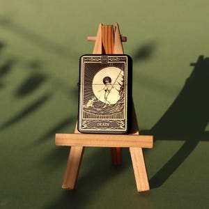 May include: A black lighter with a gold design featuring the Death tarot card. The card depicts a skeleton holding a scythe and standing on a hill. The lighter is displayed on a small wooden easel.
