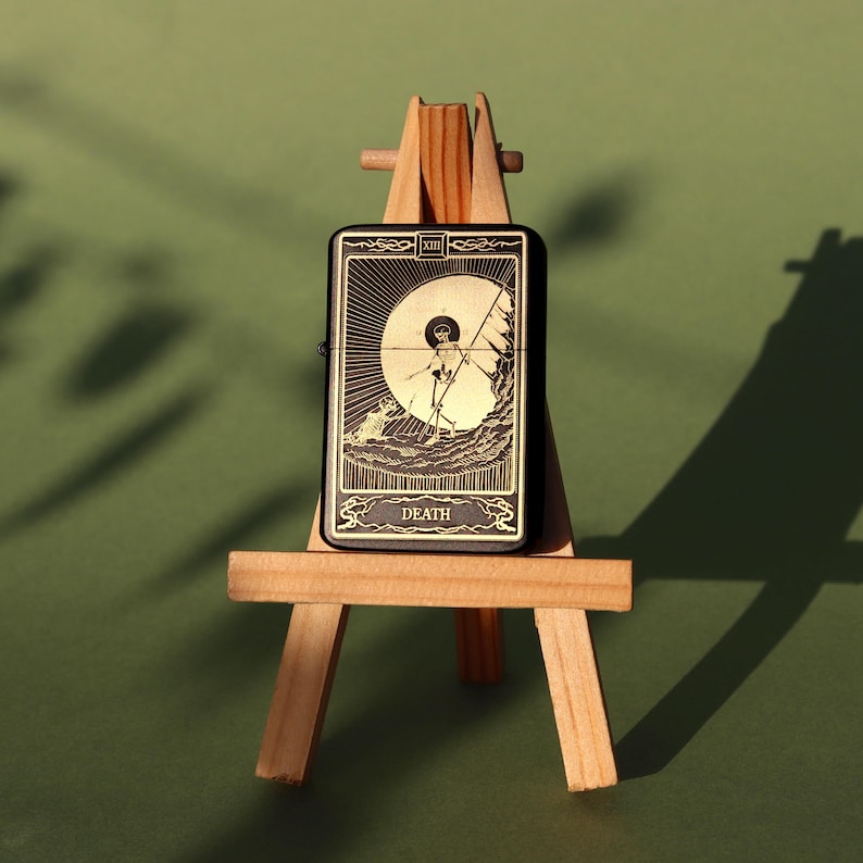 May include: A black lighter with a gold design featuring the Death tarot card. The card depicts a skeleton holding a scythe and standing on a hill. The lighter is displayed on a small wooden easel.