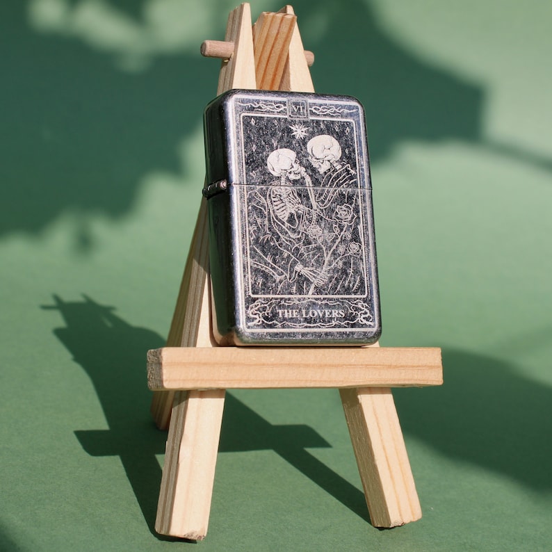 May include: A silver lighter with a black and white image of two skeletons embracing. The lighter is on a wooden easel and the text "THE LOVERS" is printed on the lighter.