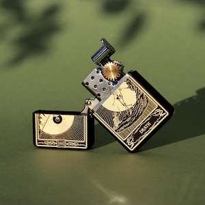 May include: A black and gold lighter with a design of the Death tarot card. The lighter has a wheel on the side and the words "Death" and "XIII" on the front.