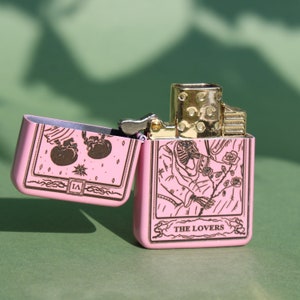 May include: Pink lighter with a gold trim and a design of the Lovers tarot card. The card features two figures holding hands and a rose.