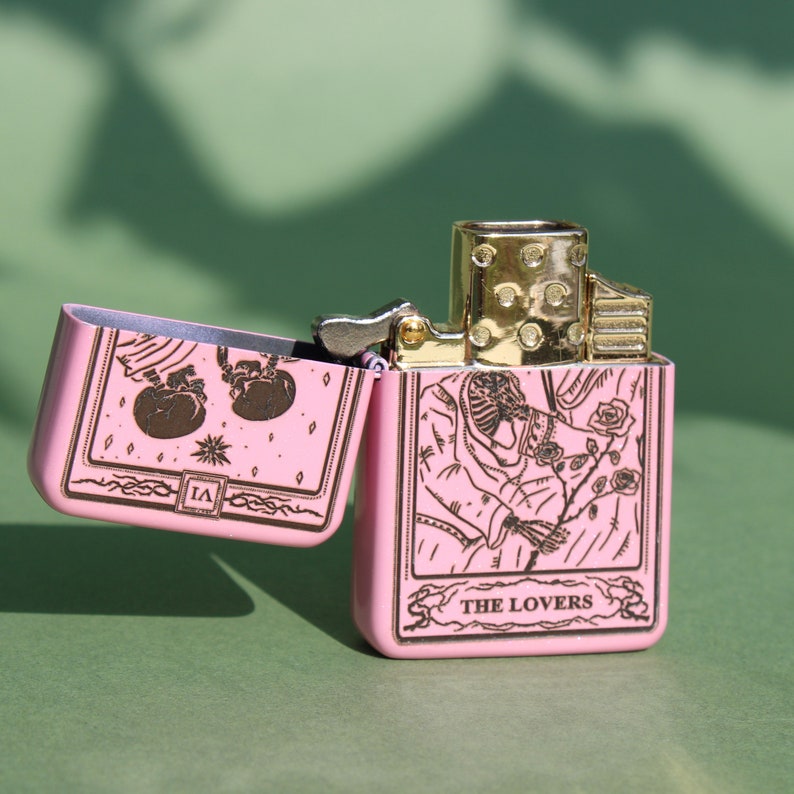 May include: Pink lighter with a gold trim and a design of the Lovers tarot card. The card features two figures holding hands and a rose.
