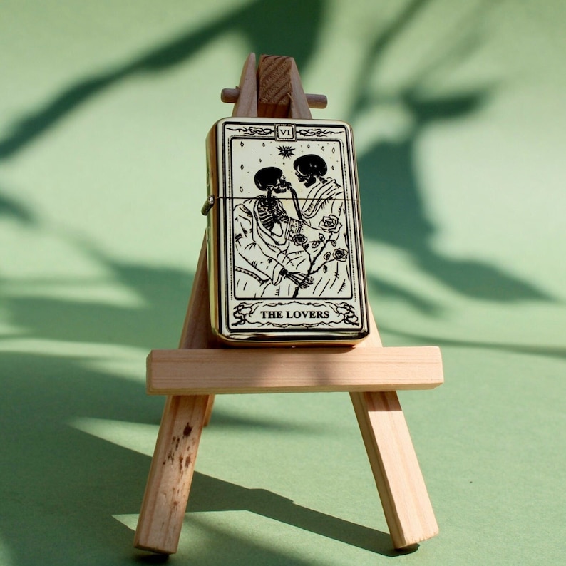 May include: A gold lighter with a black and white image of two skeletons holding a rose. The lighter is on a wooden easel and the text "The Lovers" is printed on the lighter.