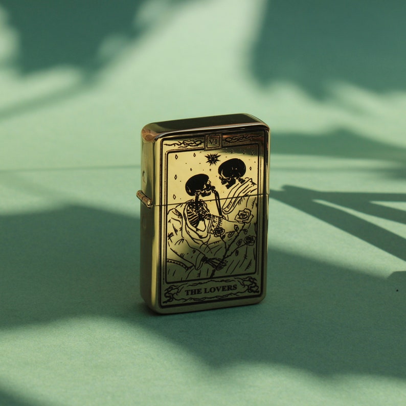 May include: A gold-colored lighter with a black and white illustration of two skeletons embracing. The lighter has the text "THE LOVERS" on the front.