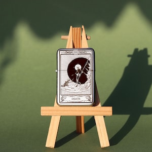 May include: A silver lighter with a black and white image of the Death tarot card. The card features a skeleton holding a scythe and standing on a hill. The lighter is on a small wooden easel.
