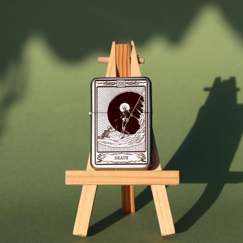May include: A silver lighter with a black and white image of the Death tarot card. The card features a skeleton holding a scythe and standing on a hill. The lighter is on a small wooden easel.