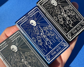 May include: Three metal lighters with a black, blue, and silver finish. Each lighter features an engraved design of a skeleton standing in a field of sunflowers with the text "THE SUN" at the bottom. The top of each lighter has the Roman numeral XIX.