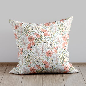 Puede incluir: Una almohada blanca con un patrón floral con flores rosas y hojas verdes. La almohada está sobre un suelo de madera.