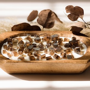 May include: A wooden dough bowl candle filled with white wax and brown crystal-like stones. The candle is surrounded by dried plants and leaves, creating a rustic and natural aesthetic.