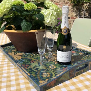 Op de afbeelding: Een rechthoekige serveerschaal met een bloemmotief in blauw, groen en goud, met twee champagneflutes en een fles Pol Roger champagne. Er staat ook een grote pot met groene hortensia's.