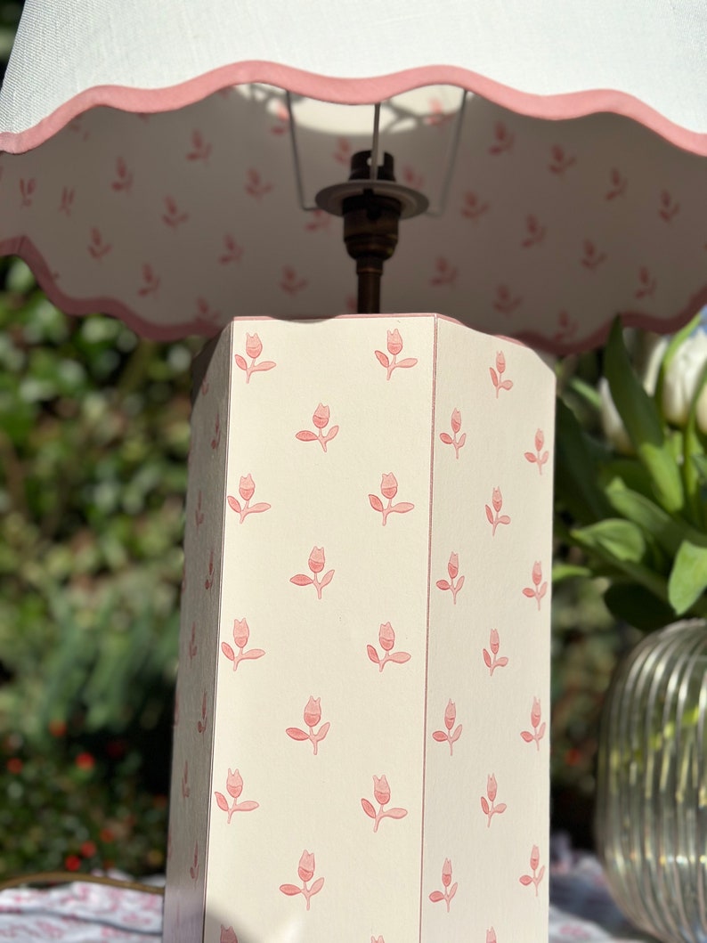 Op de afbeelding: Een witte tafellamp met een roze bloemmotief op de voet en een witte lampenkap met een roze gegolfde rand.
