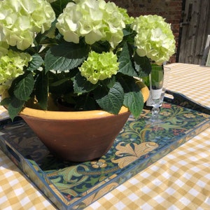 May include: A decorative wooden serving tray with a floral design in blue, green, and yellow sits on a yellow and white checkered tablecloth. A large brown bowl with a yellow rim holds green and white hydrangeas. A champagne glass is on the tray.