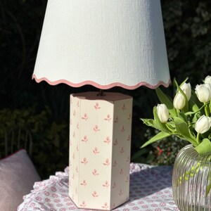 Op de afbeelding: Een roze en witte zeshoekige tafellamp met een witte linnen lampenkap met een roze gegolfde rand. De lampenvoet heeft een herhalend patroon van roze tulpen.