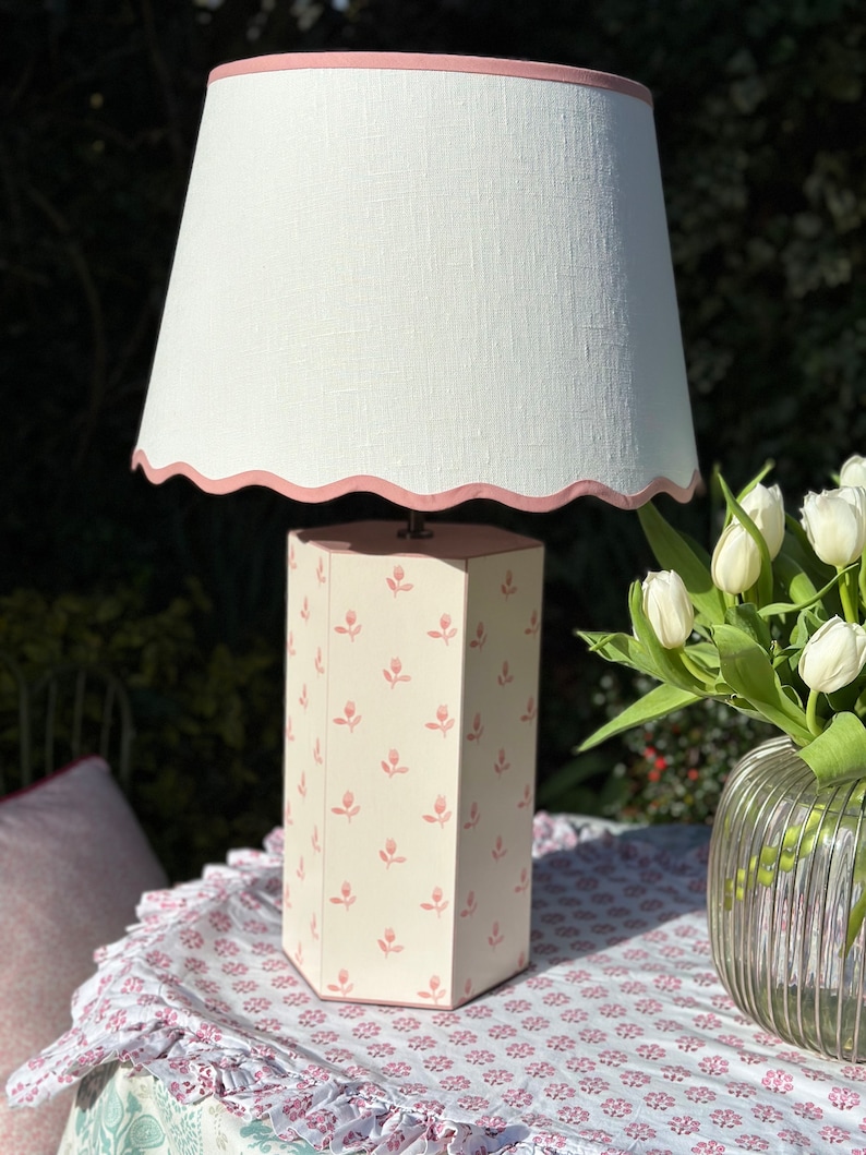 Op de afbeelding: Een roze en witte zeshoekige tafellamp met een witte linnen lampenkap met een roze gegolfde rand. De lampenvoet heeft een herhalend patroon van roze tulpen.
