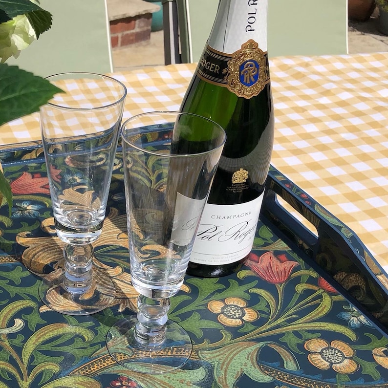 May include: A bottle of Pol Roger champagne and two clear champagne flutes on a decorative tray with a floral pattern. The tray is on a yellow and white checkered tablecloth. The champagne bottle has a gold and blue label.