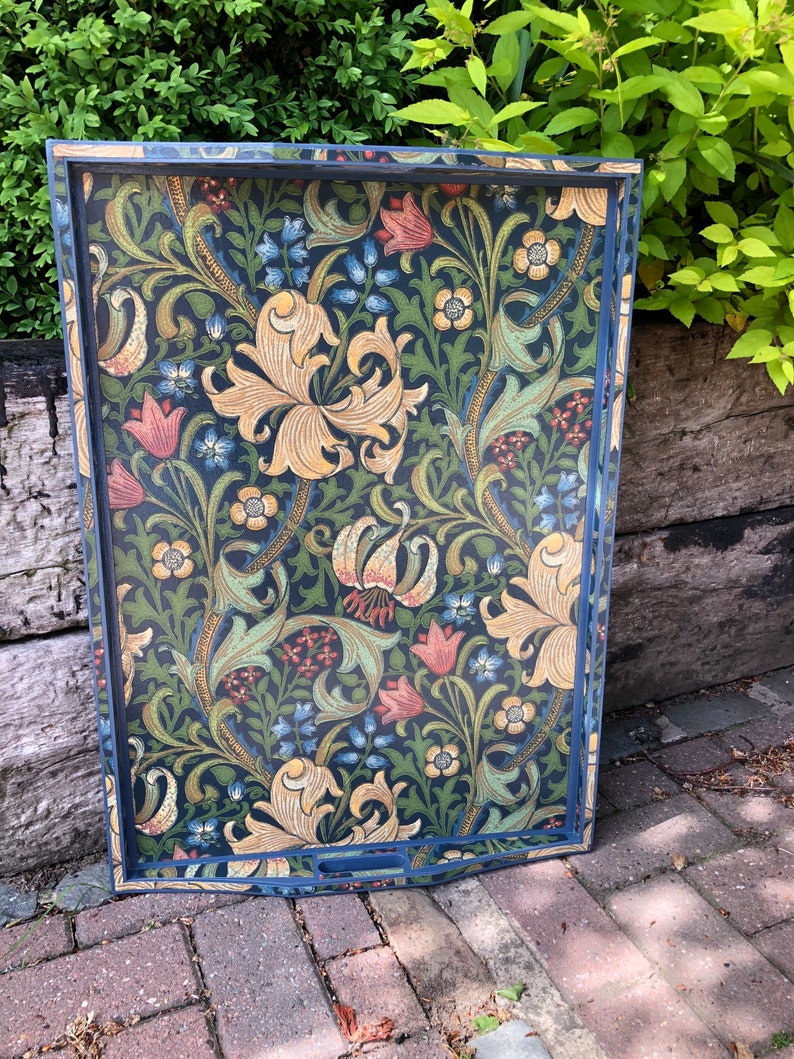 May include: Decorative serving tray with a dark blue frame and a floral pattern. The pattern features large yellow lilies, red and blue flowers, and green foliage on a dark background. The tray has a rectangular shape with curved handles.