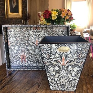 May include: A black and white tray and wastebasket with a floral pattern. The pattern features stylised flowers, leaves, and stems. The tray and wastebasket are made of wood and have a black border.