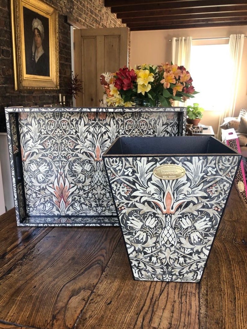 May include: A black and white tray and wastebasket with a floral pattern. The pattern features stylised flowers, leaves, and stems. The tray and wastebasket are made of wood and have a black border.