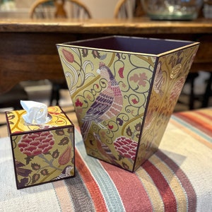 May include: A set of two decorative tissue boxes with a floral and bird pattern. The boxes are made of wood and have a yellow background with red, purple, and green accents. The larger box is a wastebasket and the smaller box is a tissue box.