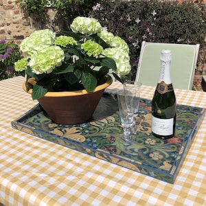 May include: A decorative serving tray with a floral design in blue, green, and gold sits on a yellow and white checkered tablecloth. A bottle of champagne and two glasses are on the tray, next to a potted hydrangea with white and green flowers.