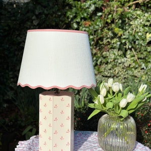 Op de afbeelding: Een witte lamp met een roze gegolfde rand op de lampenkap. De lampvoet is een witte achthoek met een roze bloemenpatroon. De lamp staat op een tafel met een roze bloemen tafelkleed.